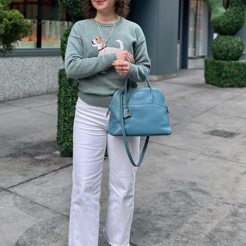 a woman in white pants and a green sweater holding a blue Hermes Bolide