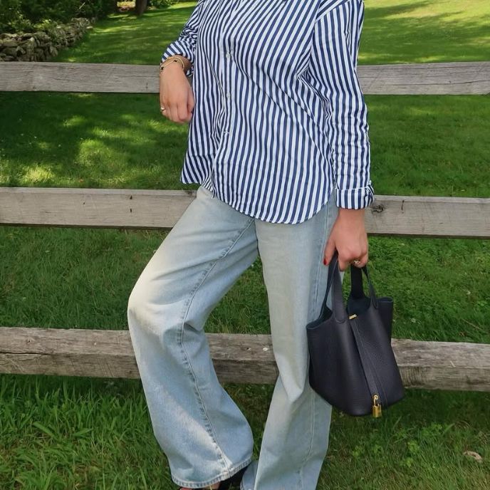 a woman in a white and navy striped button down top with denim pants holding a black Hermes Lindy bag